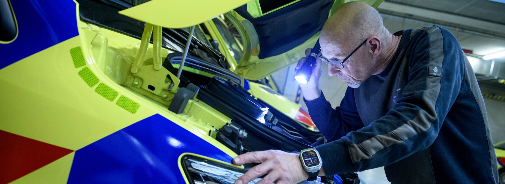 Een facilitair medewerker is met behulp van een zaklamp onder de motorkap aan het inspecteren