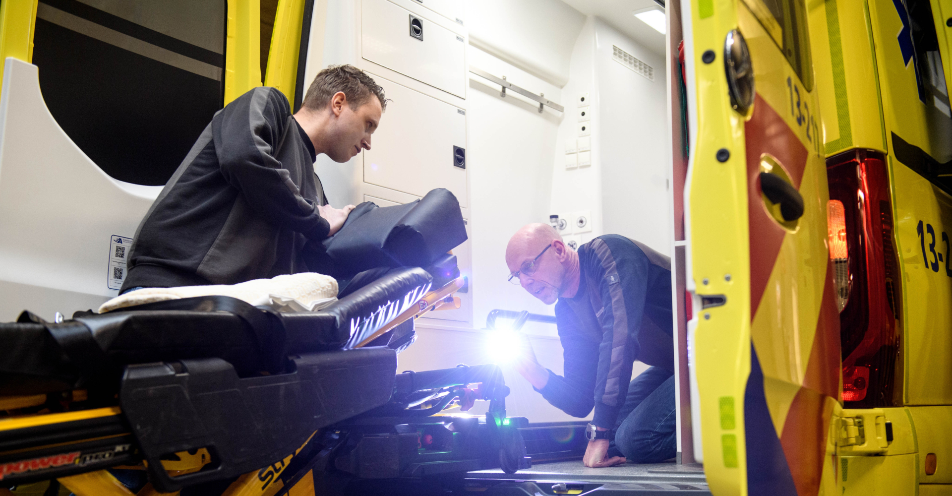 Twee facilitair medewerkers zijn aan het werk in de ambulance. Ze checken met een zaklamp de brancard