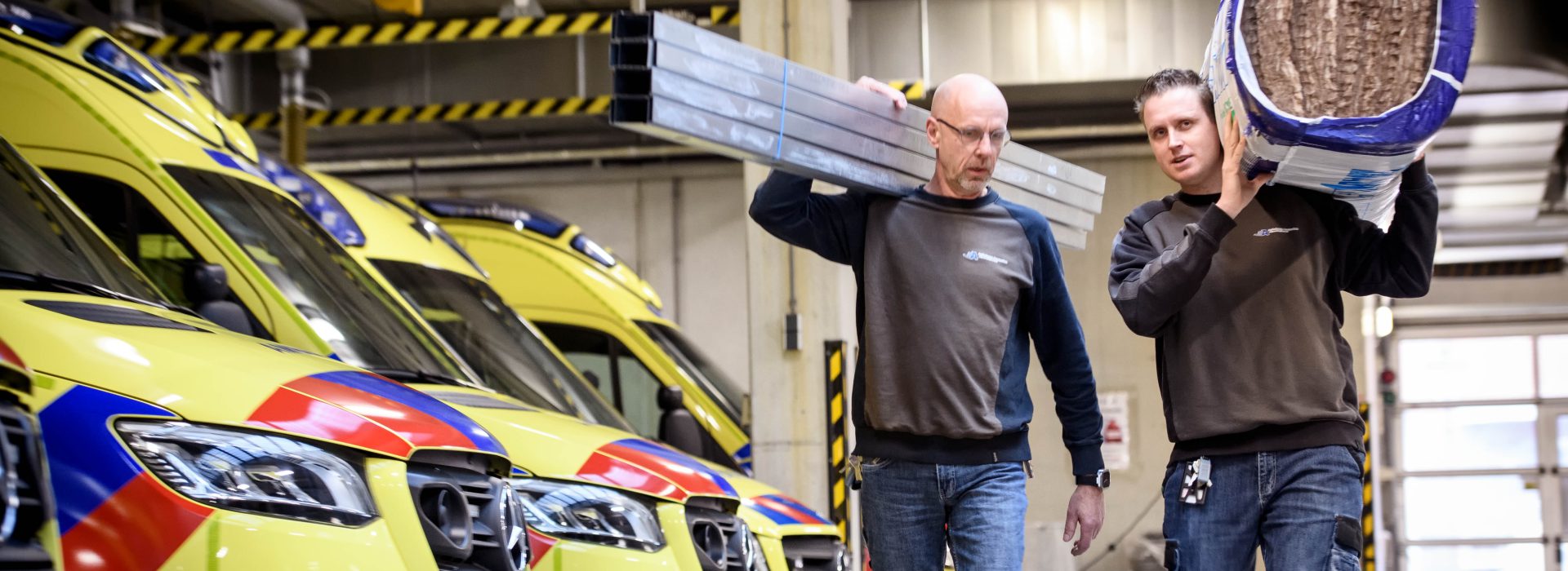 Twee facilitair medewerkers lopen met bouwmateriaal door de ambulancehal