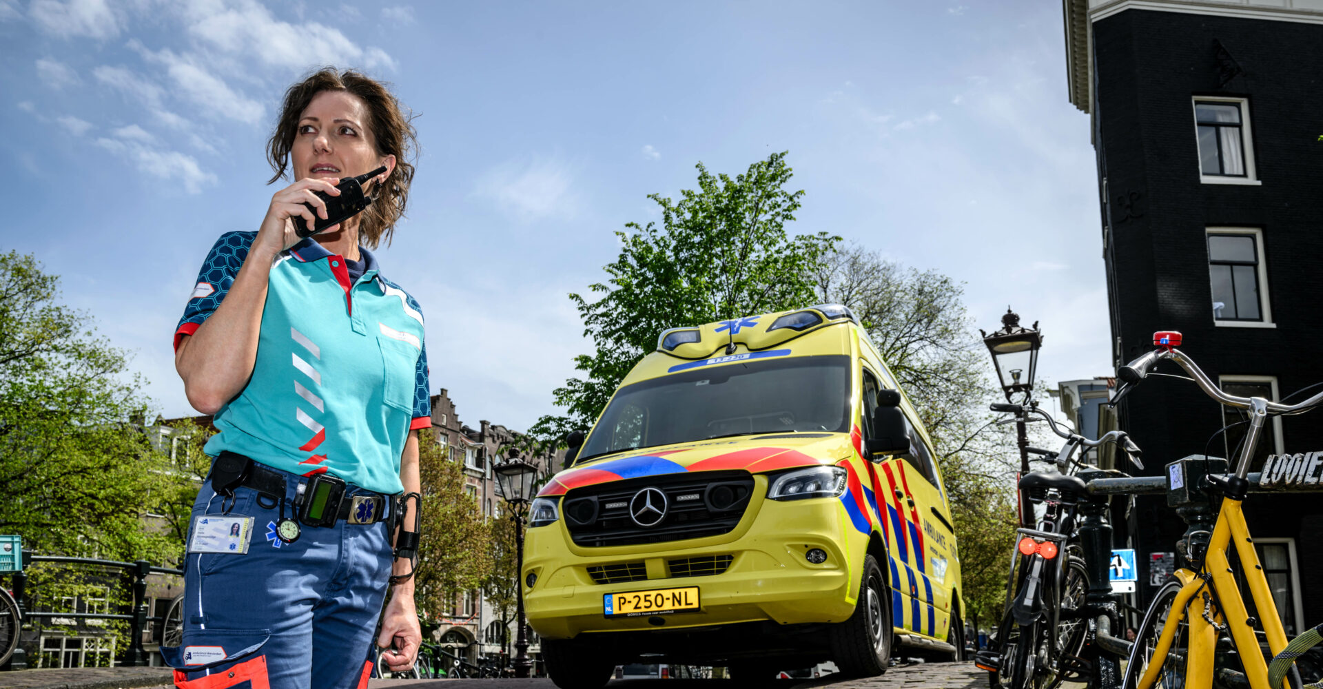 Ambulancemedewerker praat door de portofoon met de meldkamer ambulancezorg. Ze staat midden in Amsterdam, op een brug over de gracht, voor de ambulance