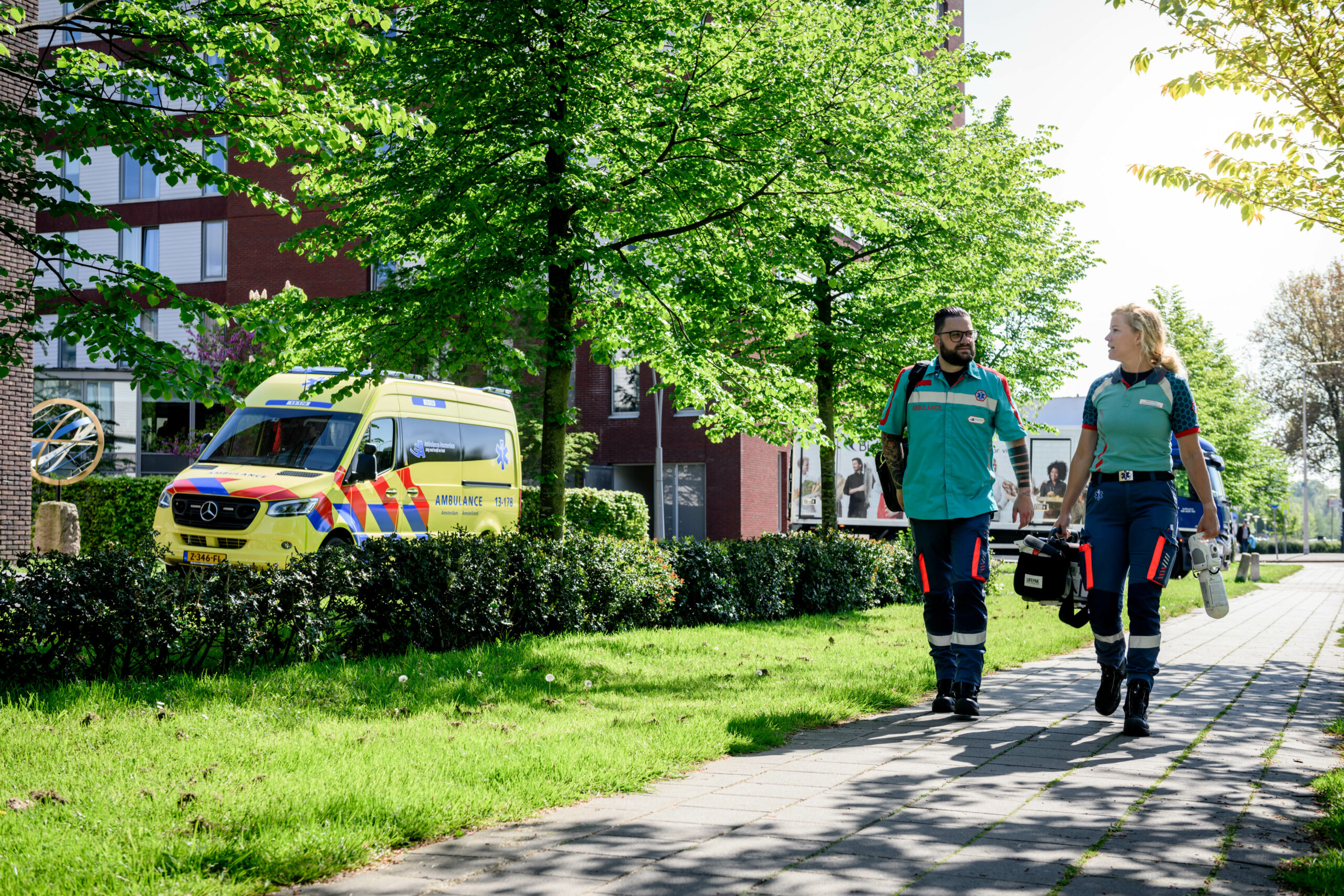 Ambulancechauffeur heeft de ambulance geparkeerd en loopt nu samen met de ambulanceverpleegkundige naar de patiënt. Ze hebben materialen zoals een ambulancetas, zuurstof en AED bij zich.