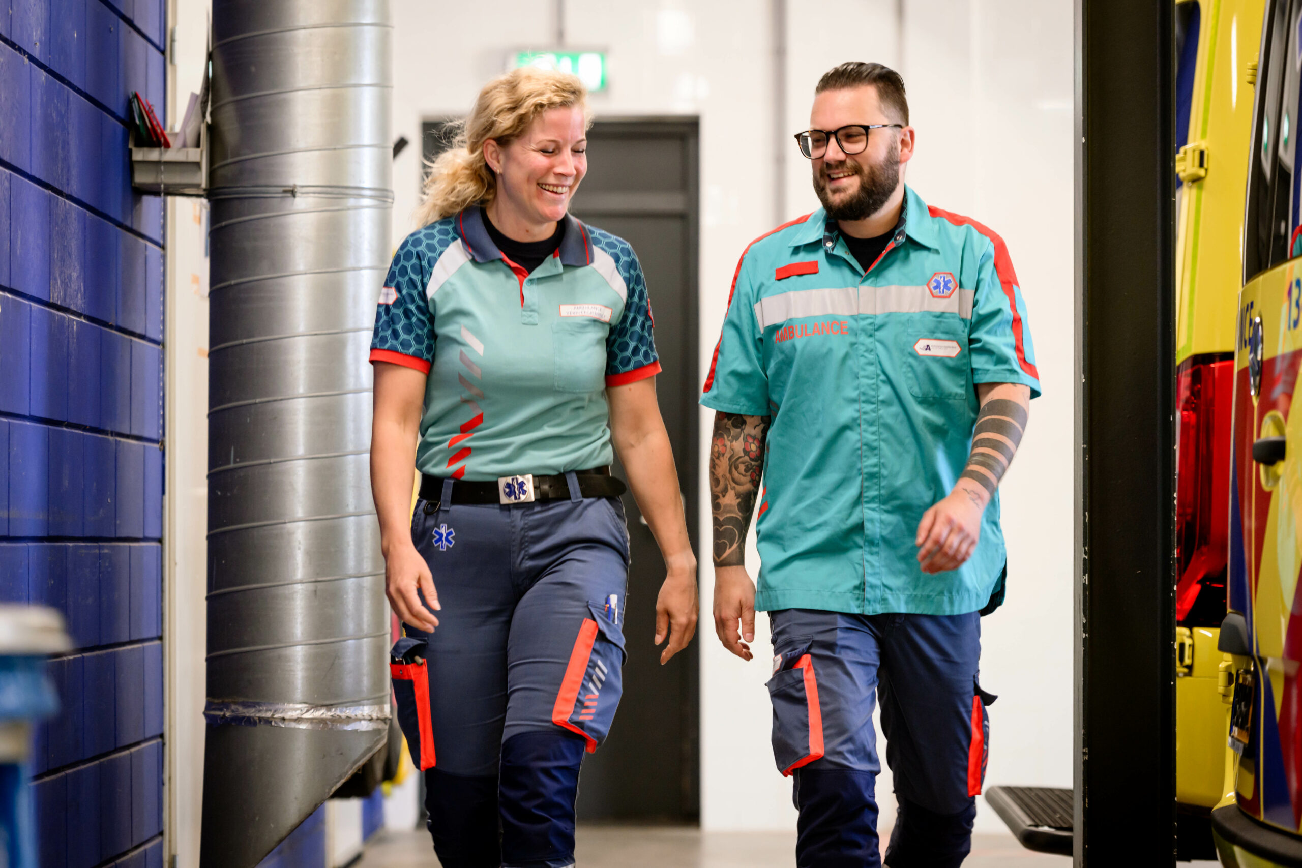Ambulanceverpleegkundige en ambulancechauffeur lopen lachend met elkaar door de ambulancehal