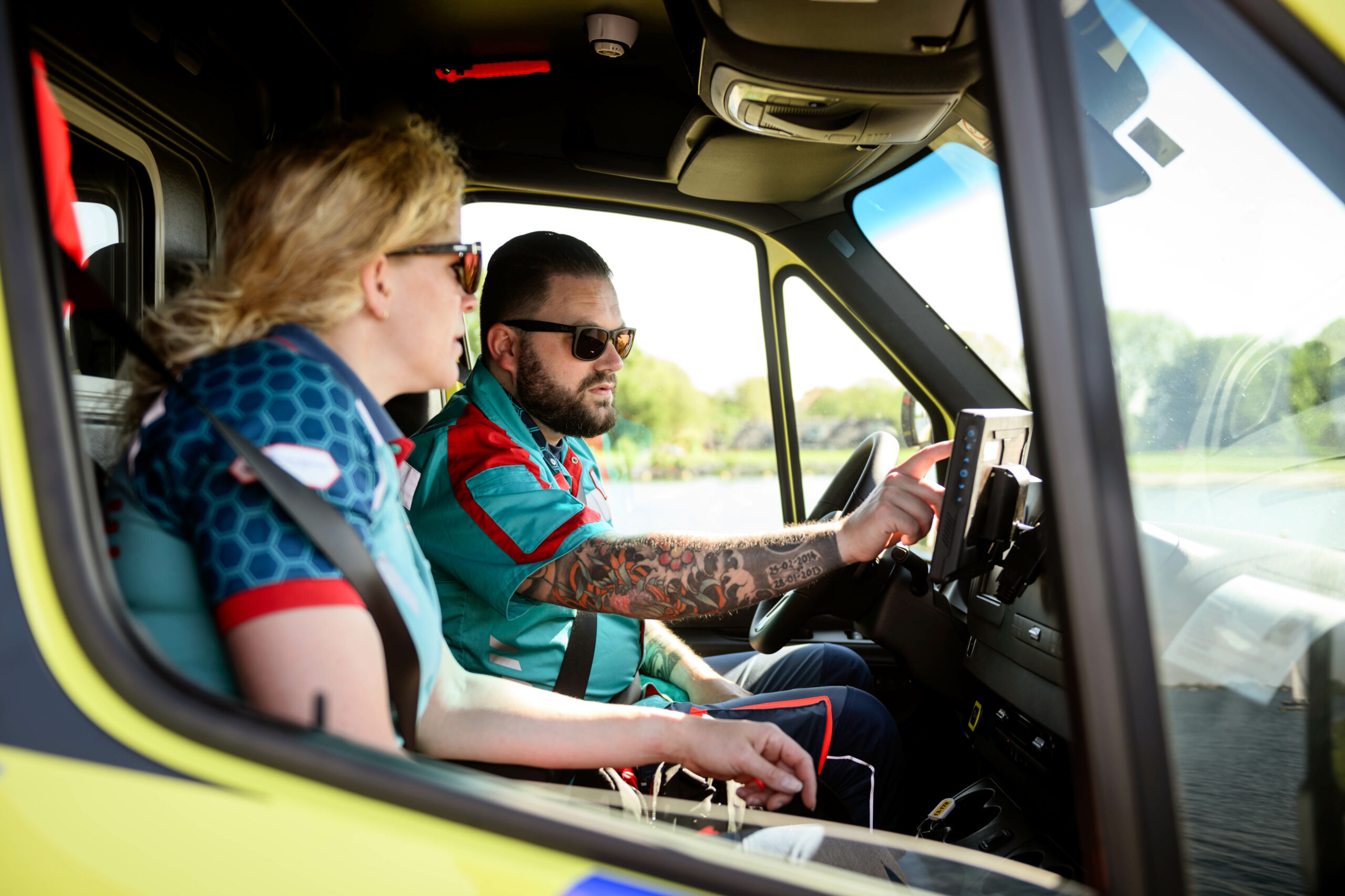 Ambulancechauffeur en ambulanceverpleegkundige zitten voorin de ambulance. De ambulancechauffeur is bezig met de tablet en de ambulanceverpleegkundige kijkt mee