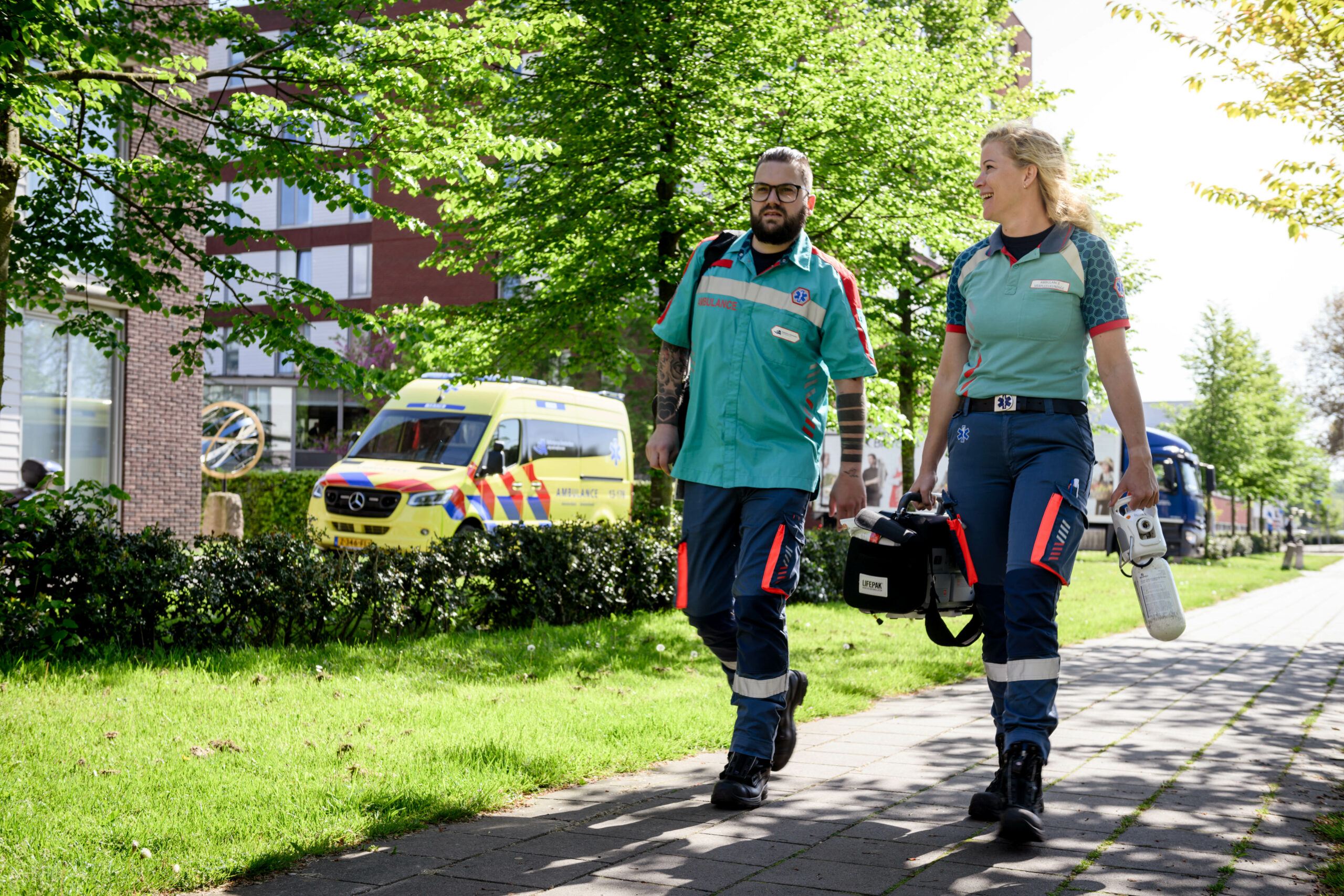 Ambulancechauffeur heeft de ambulance geparkeerd en loopt nu samen met de ambulanceverpleegkundige naar de patiënt. Ze hebben materialen zoals een ambulancetas, zuurstof en AED bij zich.