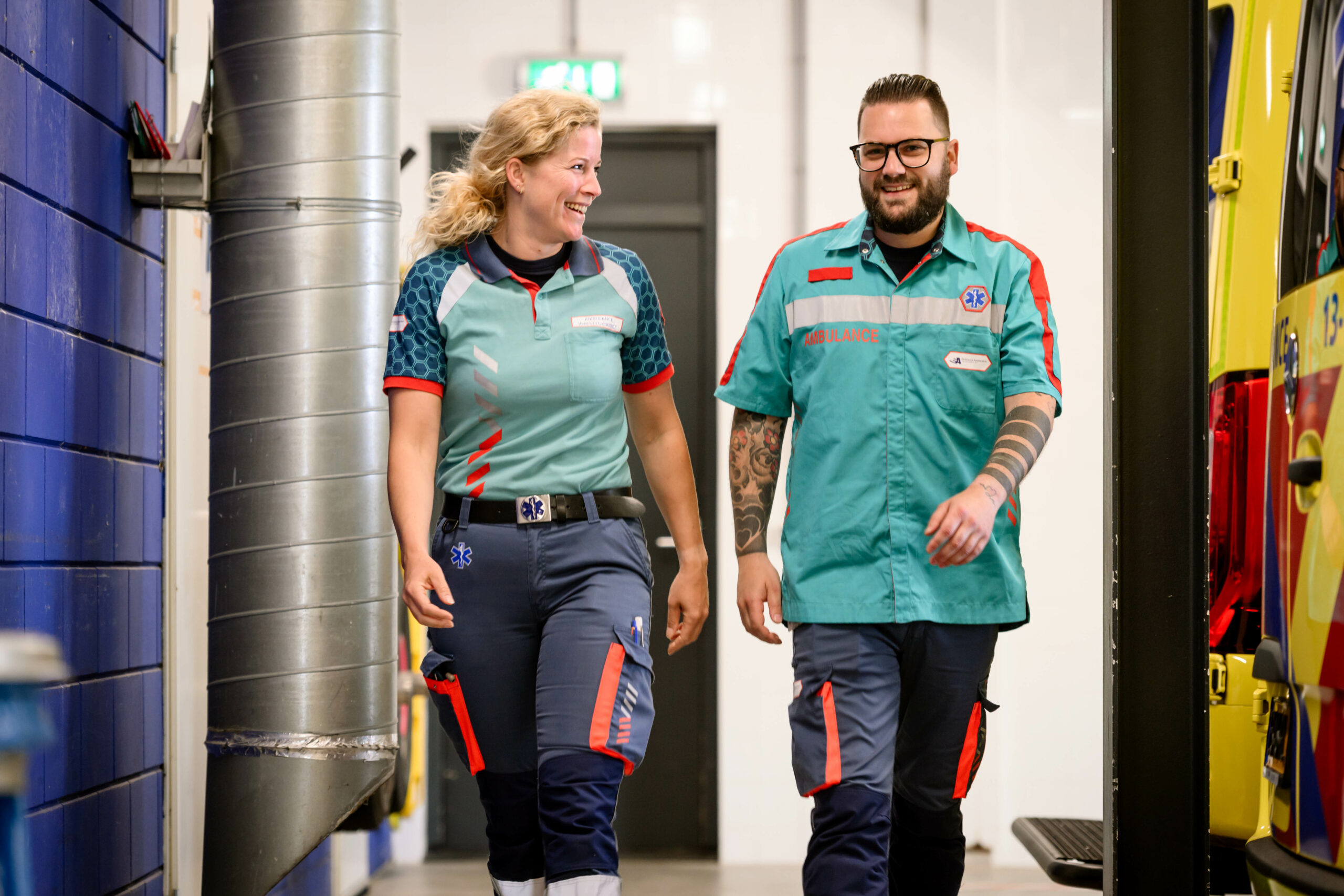 Ambulanceverpleegkundige en ambulancechauffeur lopen lachend met elkaar door de ambulancehal