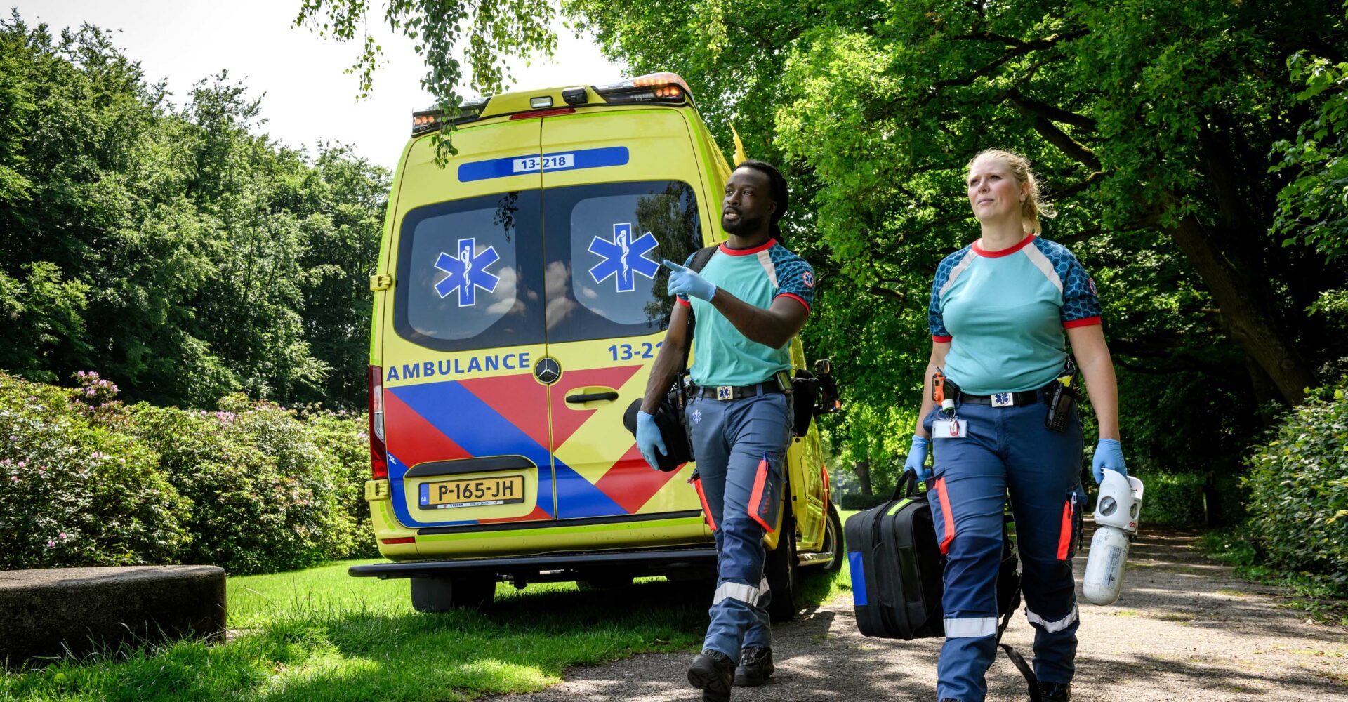 Verpleegkundige en chauffeur middencomplexe ambulance lopen in het bos richting een patiënt, met op de achtergrond een geparkeerde ambulance. Ze hebben materialen zoals zuurstof en een AED bij zich.