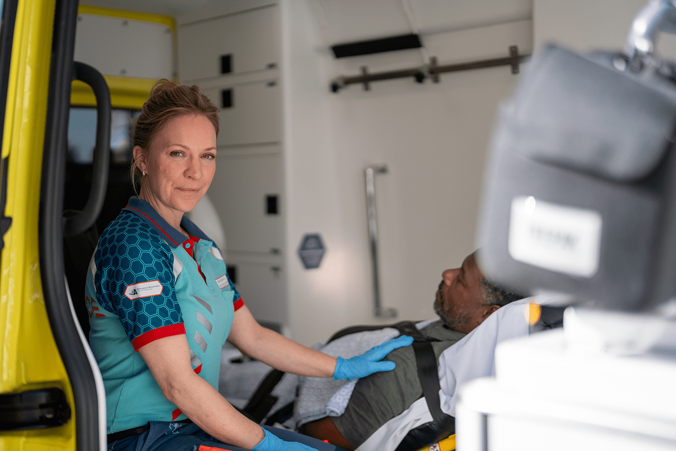 Ambulanceverpleegkundige zit in de ambulance naast een patiënt die op de brancard ligt. Ze heeft haar hand op de schouder van de patiënt