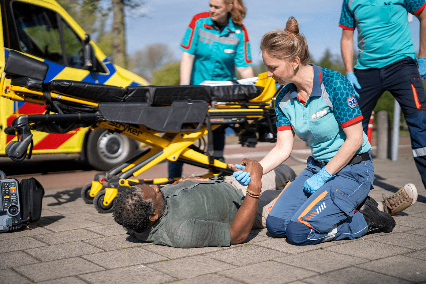 Ambulancemedewerker helpt een patiënt opstaan vanaf de grond. Op de achtergrond zie je twee ambulancemedewerkers aan het werk met de brancard.